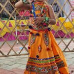 A young girl in an ornate orange lehenga stands proudly at a festive celebration.